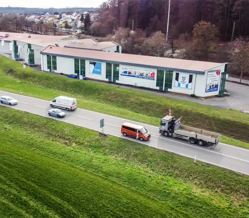 Luftaufnahme von Fahrzeugen auf einer Straße neben Industriegebäuden und Gewerbeimmobilien, angrenzend an eine Grasfläche mit Bäumen im Hintergrund.