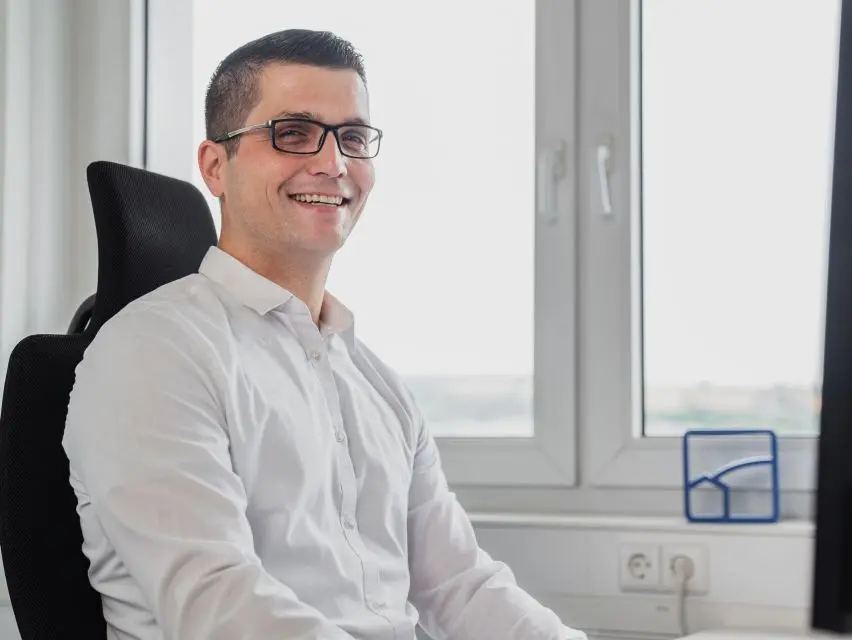 Ein Mann mit Brille und weißem Hemd sitzt lächelt auf einem Bürostuhl am Fenster und blickt auf die geschäftigen Gewerbeflächen der Rhein-Neckar-Region.