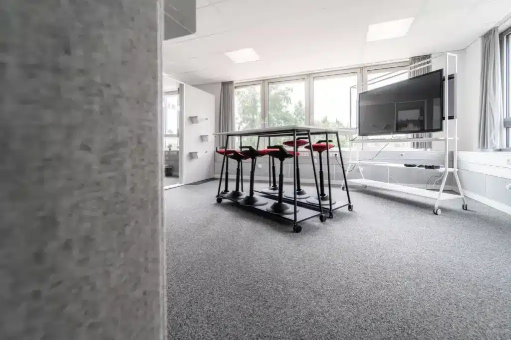 Modernes Büro mit Stehtisch, Hockern, großem Bildschirm auf Ständer, Teppichboden und großen Fenstern mit Tageslicht.