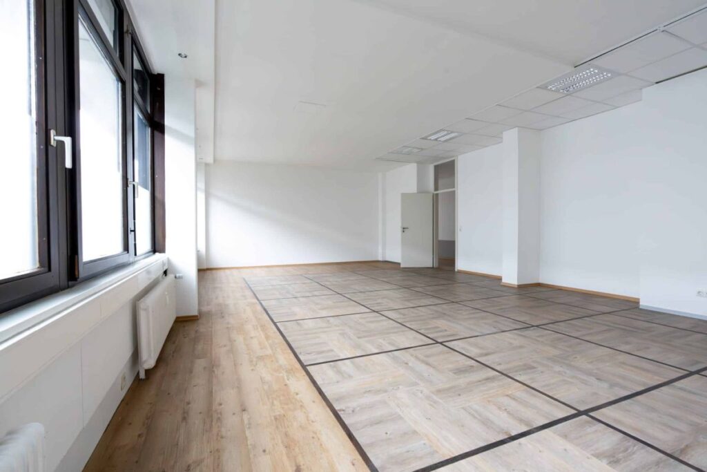 Leerer, moderner Büroraum mit großen Fenstern, weißen Wänden und hellem Holzboden mit Gittermuster.