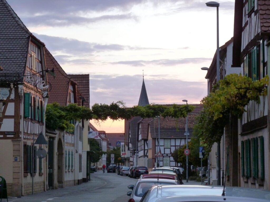 Bei Sonnenuntergang parken Autos entlang einer schmalen Straße mit traditionellen deutschen Häusern, in der Ferne ist ein Kirchturm zu sehen.