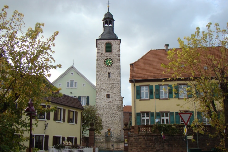 Steinturm mit grünem Ziffernblatt, umgeben von bunten Häusern und Herbstbäumen.