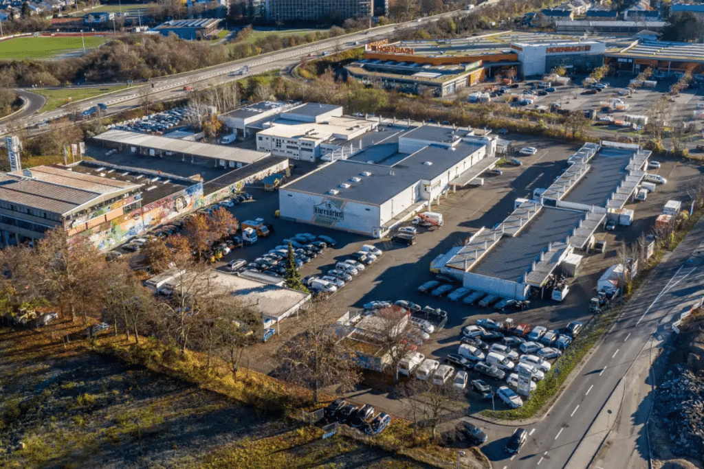 Luftaufnahme eines Industriekomplexes mit Lagerhallen, geparkten Autos sowie angrenzenden Straßen und Grünflächen.