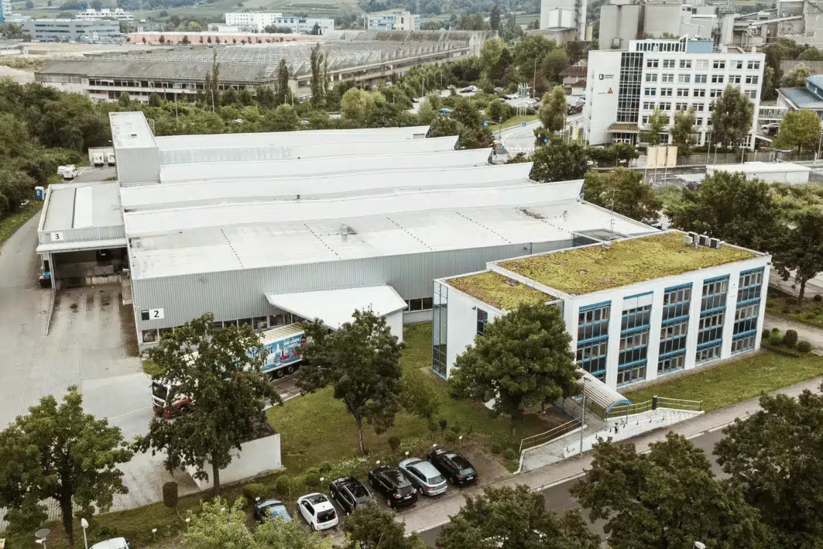 Luftaufnahme von Industriegebäuden mit begrünten Dächern und geparkten Autos, umgeben von Bäumen.