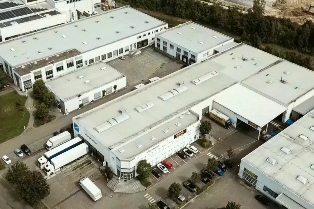 Luftaufnahme eines Industriekomplexes mit Lagerhallen, geparkten Autos, Lastwagen und umgebender Grünfläche.