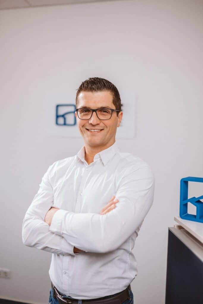 Ein Mann mit Brille und weißem Hemd steht lächelnd und mit verschränkten Armen in einem Büro.