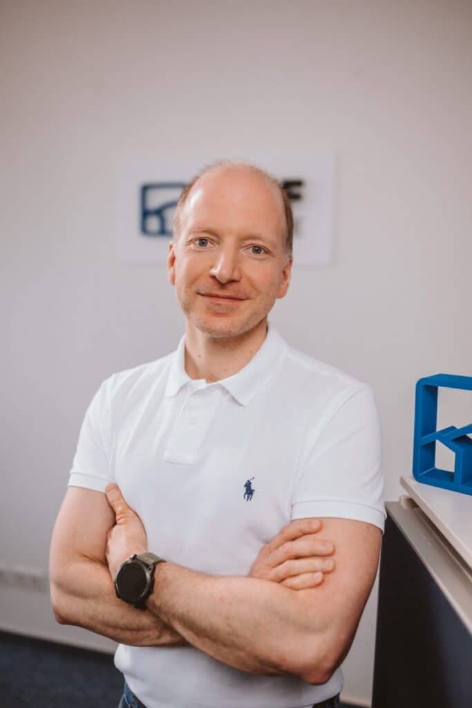 Ein Mann in einem weißen Poloshirt steht mit verschränkten Armen lächelnd in einem Büro.