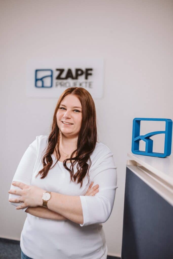 Eine Frau mit langen braunen Haaren und einem weißen Oberteil steht lächelnd neben einem blauen Logo und einem Schild mit der Aufschrift „ZAPF PROJEKTE“.