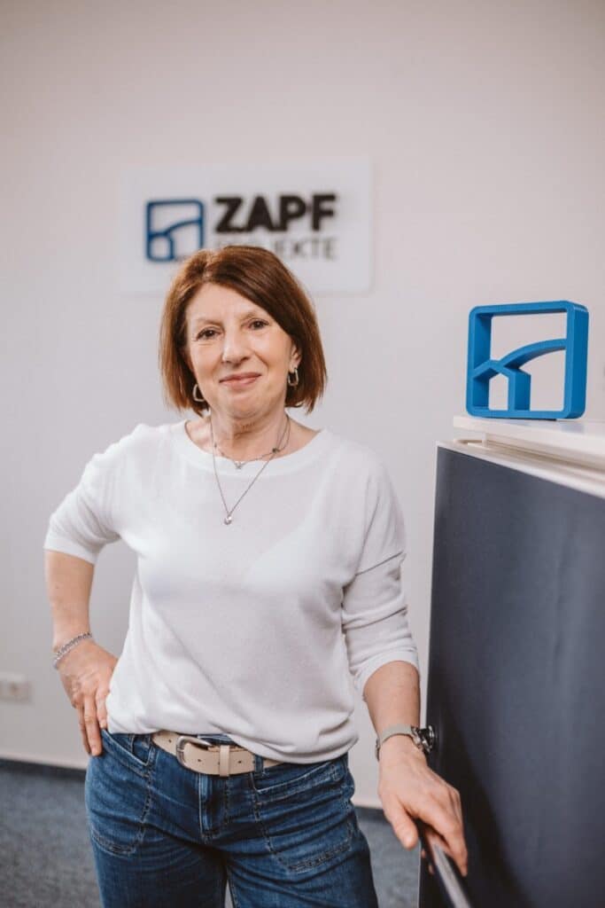 Eine Frau in einem weißen Hemd steht lächelnd in einem Büro, im Hintergrund ist ein Schild mit der Aufschrift „ZAPF PROJEKTE“ zu sehen.