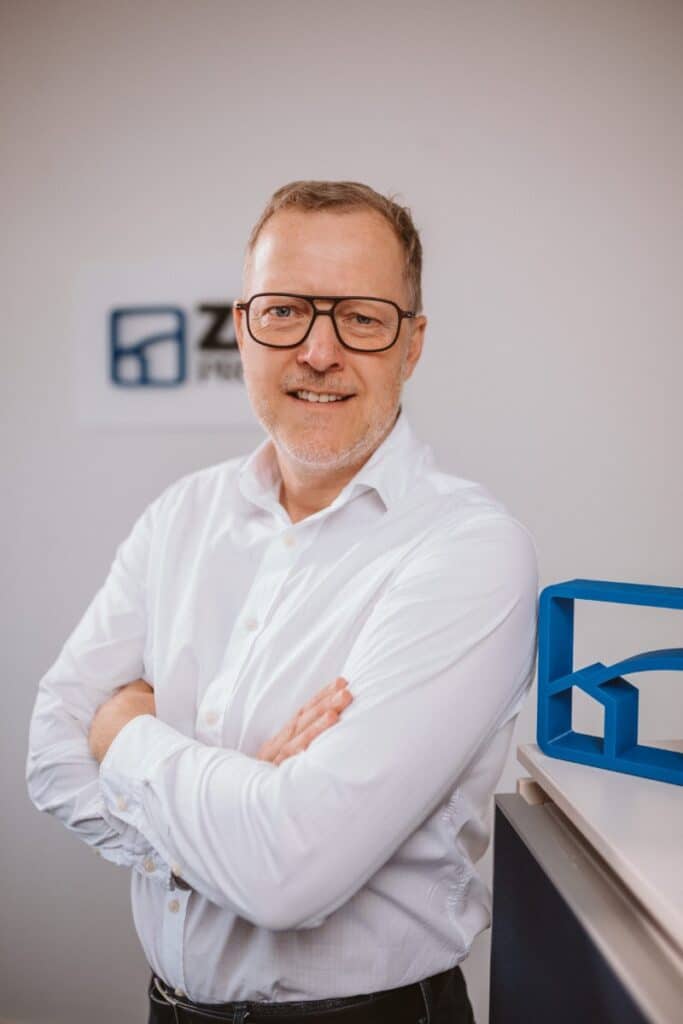 Ein Mann mit Brille und weißem Hemd, der mit verschränkten Armen lächelt und in einem Büro steht, im Hintergrund ist ein blaues Logo zu sehen.