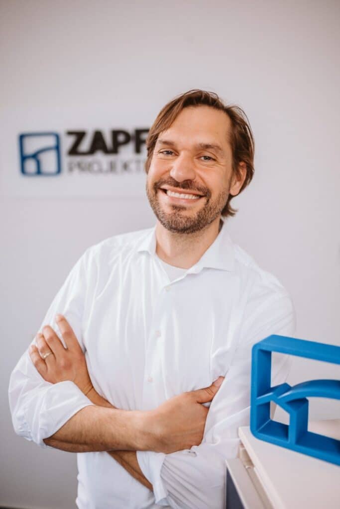 Ein Mann in einem weißen Hemd, der mit verschränkten Armen lächelt und in einem Büro steht, hinter ihm ein Schild mit der Aufschrift „Zapf Projekte“.