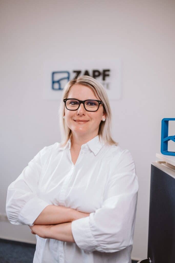 Eine Frau mit Brille und weißem Hemd steht lächelnd mit verschränkten Armen in einem Büro.