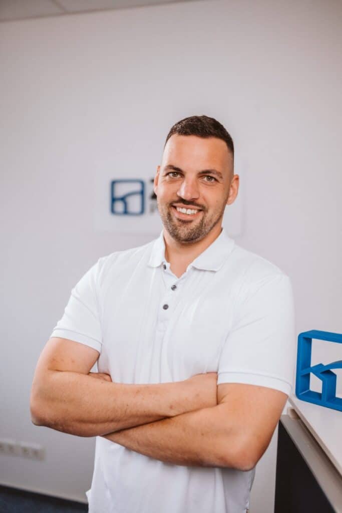 Ein Mann in einem weißen Poloshirt steht lächelnd mit verschränkten Armen in einer modernen Büroumgebung.