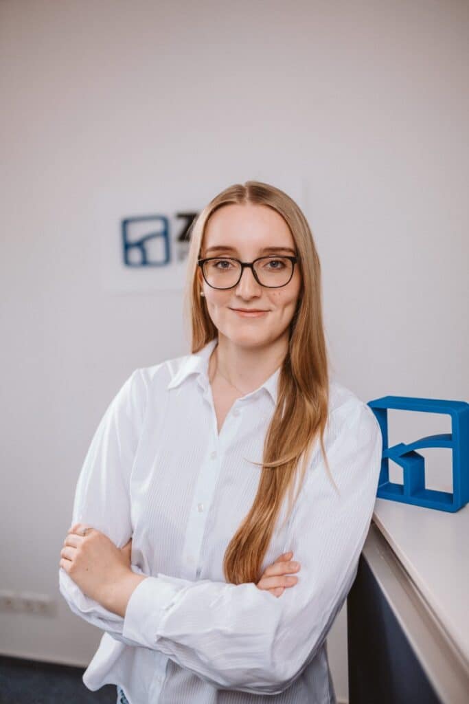 Eine Frau mit langen blonden Haaren und Brille steht lächelnd mit verschränkten Armen in einem Büro.