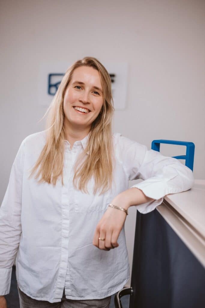 Eine Frau mit langen blonden Haaren lächelt, während sie sich in einem Büro an eine Theke lehnt; sie trägt ein weißes Hemd.