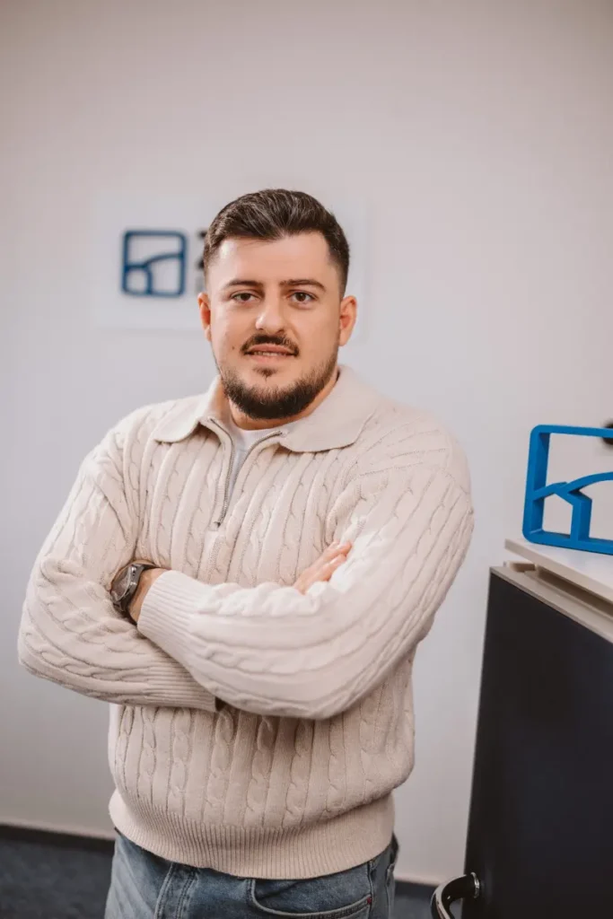 Ein Mann in einem beigen Pullover steht mit verschränkten Armen lächelnd in einem Büro mit blauen Akzenten.
