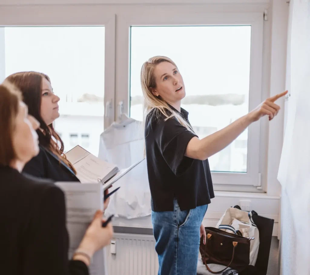 Drei Frauen in einem Büro: Eine zeigt auf ein Whiteboard, während die anderen zusehen und sich Notizen machen.